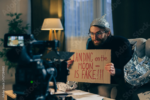 A conspiracy theorist shoots pseudoscientific videos on camera. A man in a tinfoil hat and a sign in his hands sits on a couch in front of the camera. photo