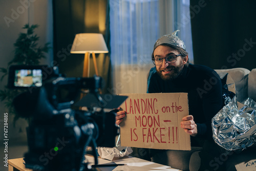 A conspiracy theorist shoots pseudoscientific videos on camera. A man in a tinfoil hat and a sign in his hands sits on a couch in front of the camera. photo