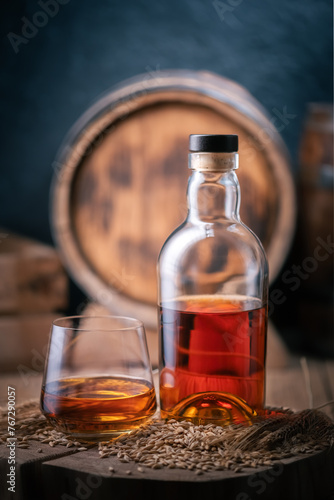 A glass of whiskey with bottle on wooden board. Oak barrel on background. Barley grains and barley ears on a foreground