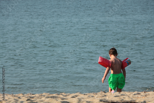 blonde boy walking towards the sea on the beach. boy in green shorts with red inflatable water armbands. Rear view of child with armbands walking on beach on summer holiday.
