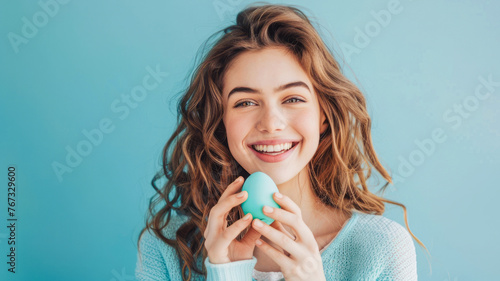 A girl smiles while holding an Easter egg, set against a light blue background, encapsulating joy and the festive spirit succinctly. photo
