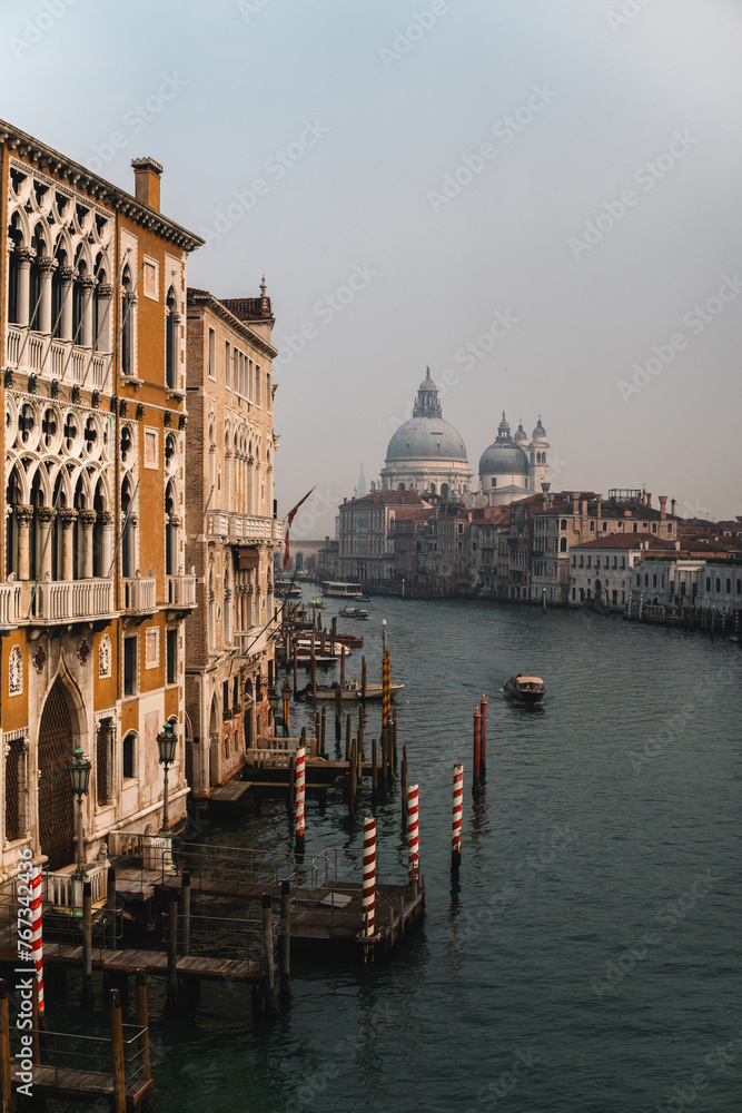 Grand Canal from Accademia Bridge: Buildings with medieval-style balconies and windows leading to wooden docks with iconic striped poles. Distant views of Santa Maria della Salute and boats cruising.