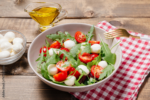 Fresh summer vegetable salad with mozzarella cheese, green arugula leaves and cherry tomatoes on wooden table background. Healthy food, diet concept. Flat lay, top view.