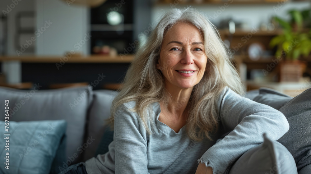 A woman with long gray hair is sitting on a couch and smiling. She is wearing a gray shirt and a gray sweater A woman with long gray hair is sitting on a couch and smiling. She is wearing a gray shirt and a gray sweater