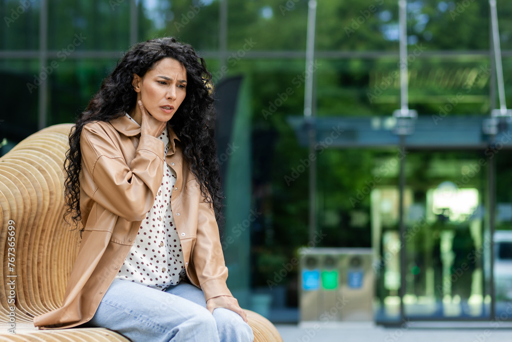 Disappointed business lady sitting on bench outside building and experiencing throat pain. Frustrated caucasian female in elegant clothes touching neck and thinking how to relieve ache. Copy space.