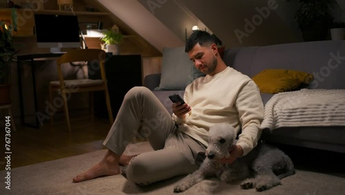 Young man holding mobile phone while sitting on carpet with dog and caresses his pet photo