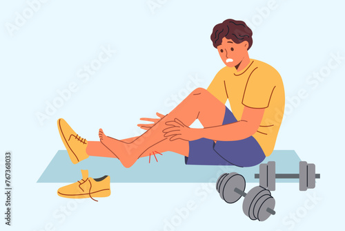 Injured sportsman sits on mat for sports and gymnastics near dumbbells, after stretching muscles on legs. Injured athlete experiences pain and agony after careless use of sports equipment
