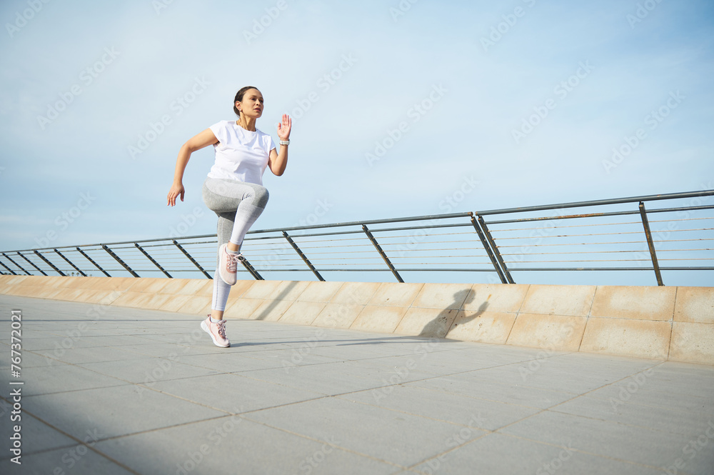Young athletic woman, jogger, runner practicing sport cardio workout outdoors, running fast along the promenade bridge over the sky and ocean background. Healthy lifestyle. Workout jogging activity