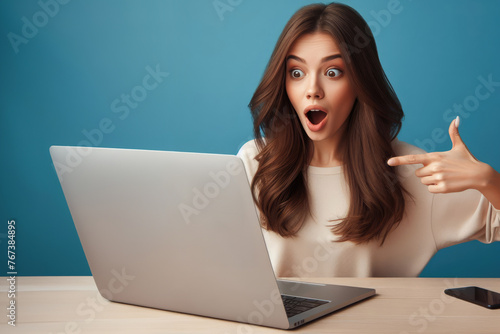 young woman working with laptop, with surprised facial expression, she sees great discounts and irresistible offers at her favorite stores, pointing to against laptop blue background.