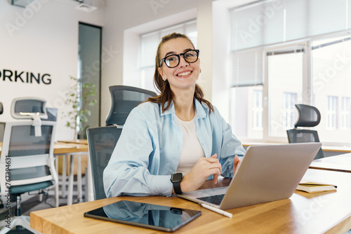 Elegantly poised at a modern workspace, a jovial young woman engages with her laptop, exuding professionalism and contentment amidst a well-lit office.