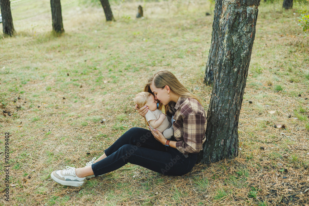 A cute mother is sitting in the park on the grass with a small child in her arms. A walk with a small child in nature. A cute mother is sitting in the park on the grass with a small child in her arms. A walk with a small child in nature.