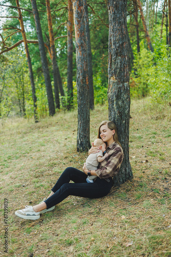 A cute mother is sitting in the park on the grass with a small child in her arms. A walk with a small child in nature.