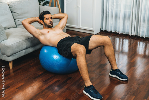 Athletic and sporty man doing situp on fitness ball during home body workout exercise session for fit physique and healthy sport lifestyle at home. Gaiety home exercise workout training concept.