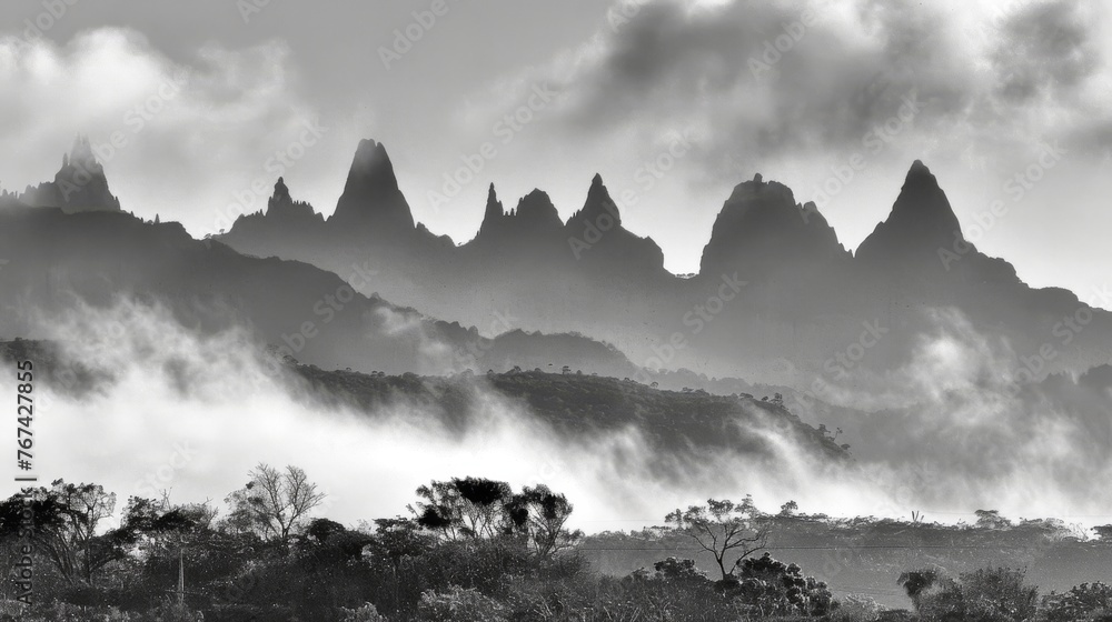  a black and white photo of a mountain range with low clouds in the foreground and trees in the foreground.