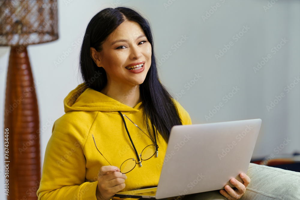 A woman in a yellow hoodie is smiling and holding a laptop. She is wearing glasses and she is enjoying her time A woman in a yellow hoodie is smiling and holding a laptop. She is wearing glasses and she is enjoying her time