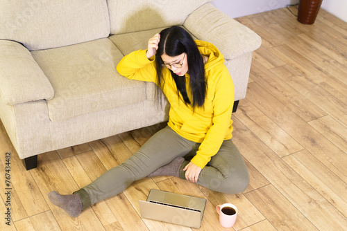 A woman is sitting on the floor with a laptop in front of her. She is wearing a yellow hoodie and has glasses on. The scene is casual and relaxed