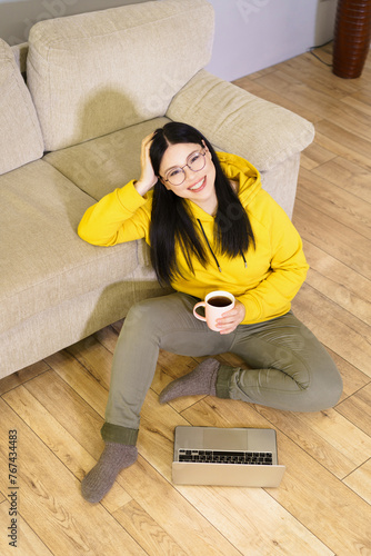 A woman in a yellow hoodie is sitting on the floor with a laptop and a cup of coffee. She is smiling and she is enjoying her time