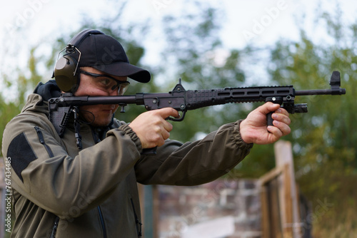 A man is holding a gun and wearing a green jacket. He is wearing a black hat and glasses