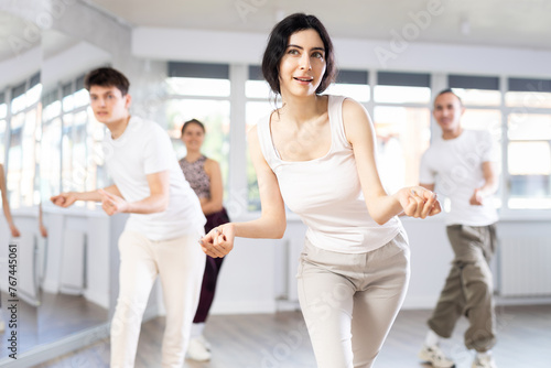 Smiling young dark-haired woman rehearsing jazz-style routine in dance studio, performing traveling steps and snapping fingers