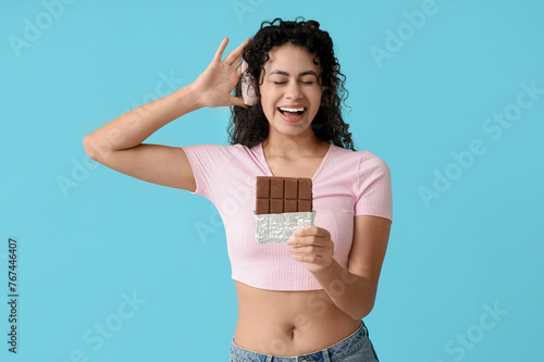 Beautiful young African-American woman in headphones with sweet chocolate bar on blue background © Pixel-Shot