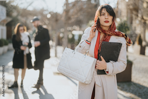 Elegant young woman in a chic coat and stylish scarf holding a handbag and folder, walking on a sunny sidewalk in the city.