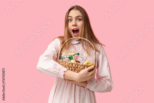 Surprised young woman holding basket with makeup products, flowers and Easter decor on pink background