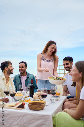 Vertical. Group of people gathered around table, sharing food and wine while smiling and enjoying leisurely event. The hostess serves food to her guests. Friends lunch together on a spring summer day