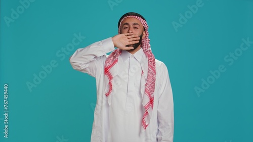 Sleep deprived guy yawning in studio, feeling extremely sleepy and almost falling asleep standing on camera. Islamic person with thobe and kufiyah being exhausted, suffering from fatigue. photo