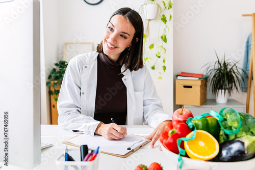 Young female nutritionist talking with patient through desktop computer video call at clinical consultation room. Healthy lifestyle, nutrition and diet concept.