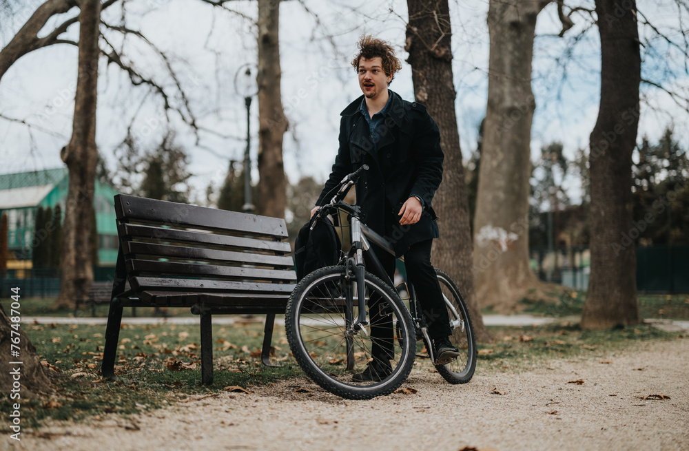 A young adult male holding a bicycle stands next to a wooden park bench, surrounded by autumn leaves.