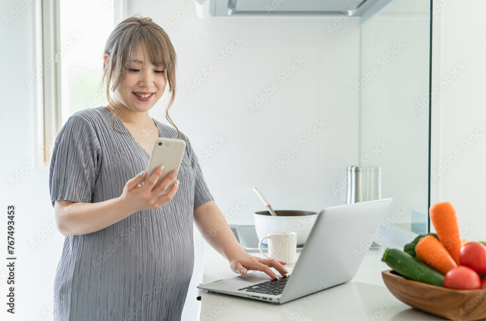 キッチンで料理をしながらパソコンとスマホを使う妊婦の女性
キッチンで料理をしながらパソコンとスマホを使う妊婦の女性