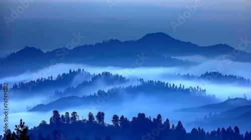  a view of a foggy mountain range with pine trees in the foreground and blue sky in the background.