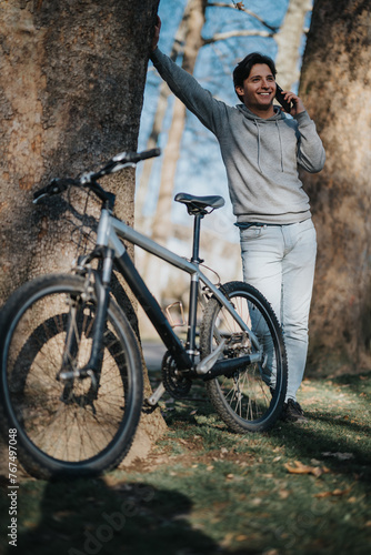 A cheerful young man enjoys a phone conversation outdoors in a park with his bicycle by his side on a sunny day.