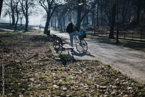 A lone individual walks with their bike on a serene path in a park, surrounded by autumn leaves and empty benches.