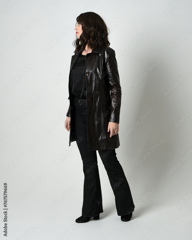 Full length portrait of brunette woman wearing long leather trench coat. Standing pose walking away from camera, looking back over shoulder, reaching out. Isolated on white studio background. Full length portrait of brunette woman wearing long leather trench coat. Standing pose walking away from camera, looking back over shoulder, reaching out. Isolated on white studio background.