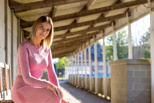 An athletic woman in pink sportswear finds a moment of quiet rest on a wooden bench under the linear canopy of an urban sports facility. Athletic Rest: Woman in Pink Sportswear Enjoying Urban Solitude