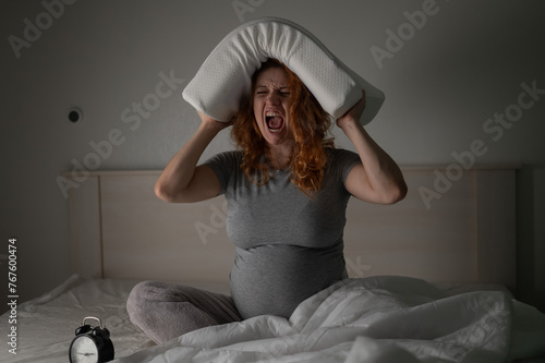 Caucasian pregnant woman sits in bed, holds a pillow on her head and screams. Insomnia. photo