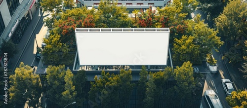 A large empty poster mockup displayed on top of a shopping center, featuring a white placeholder template of a billboard on the rooftop of a contemporary structure surrounded by trees. © Vusal