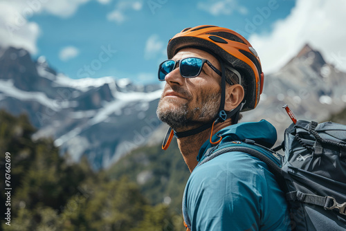 A man wearing a helmet and sunglasses is smiling. The scene is set in a mountainous area. Man wearing helmet stands confidently before towering mountain backdrop ready for adventure and exploration.