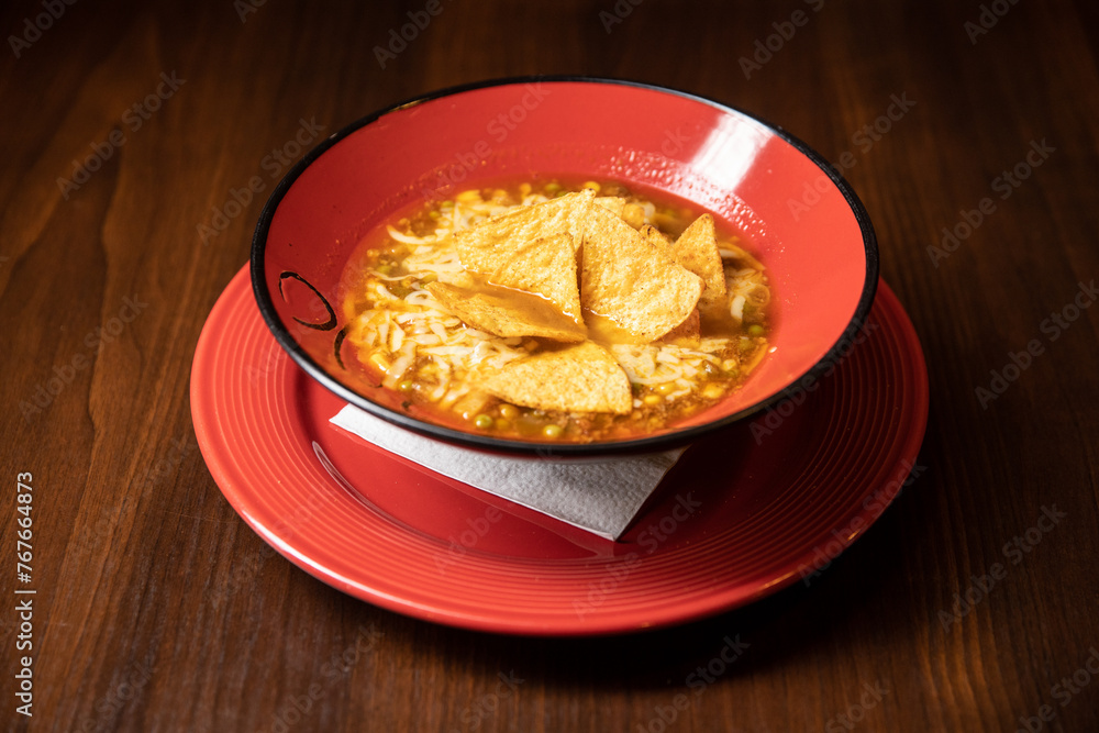 mexican tortilla soup served in red dish in restaurant with red cocktail on dark background