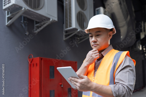 Close-up photo of a thoughtful and serious young Asian man, a factory worker, a construction worker and an engineer standing outside in a hard hat and vest, using a tablet