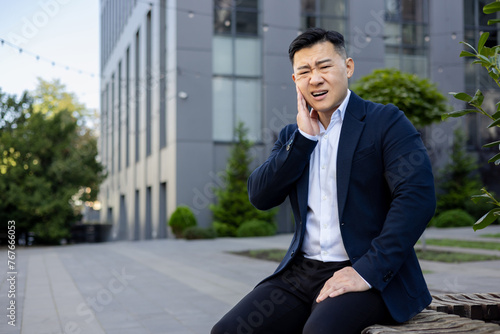 Asian young male businessman sitting on a bench outside an office building and crookedly holding his hand to his cheek, suffering from severe toothache and discomfort