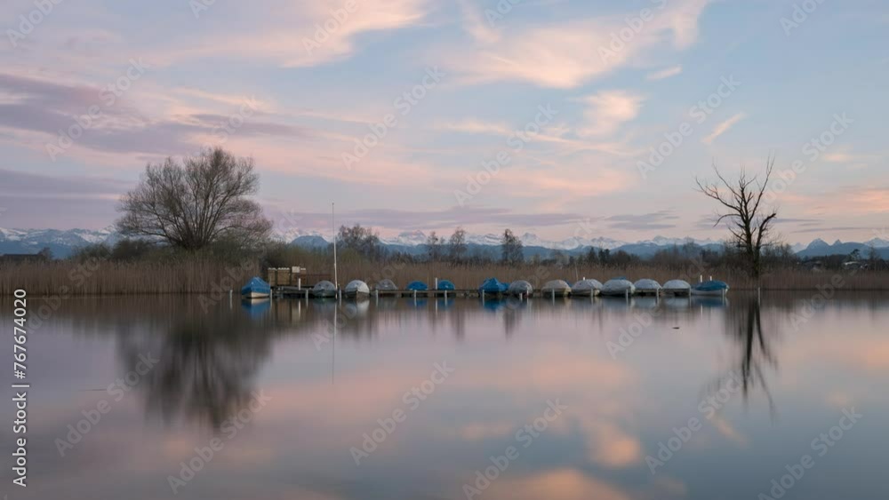 Calm sunset on a lake with snowy mountains in the background and boats on the water. The landscape reflects in the water and the sky turns pink Calm sunset on a lake with snowy mountains in the background and boats on the water. The landscape reflects in the water and the sky turns pink