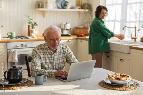 elderly grey haired man in eyeglasses uses laptop screen in kitchen photo