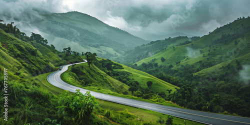 A winding road cuts through a lush, verdant landscape of rolling hills and dense forests under an overcast sky.