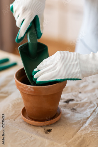 people, gardening and planting concept - close up of woman in gloves with trowel pouring soil to flower pot at home