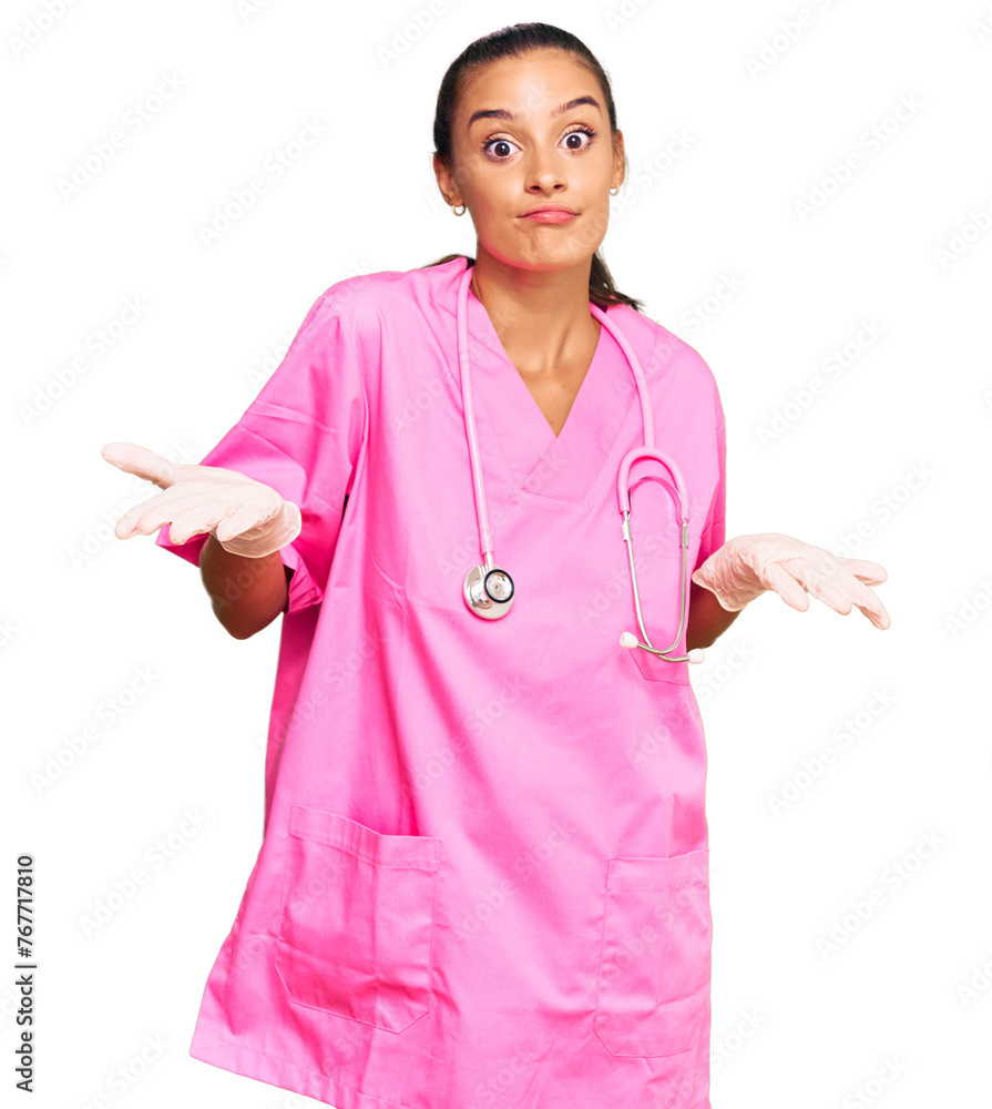 Young hispanic woman wearing doctor uniform and stethoscope clueless and confused expression with arms and hands raised. doubt concept. Young hispanic woman wearing doctor uniform and stethoscope clueless and confused expression with arms and hands raised. doubt concept.