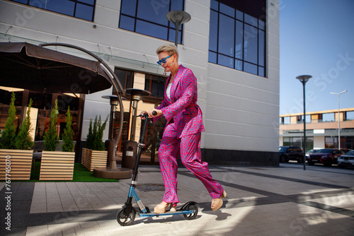 Stylishly dressed mature woman in a bright pink pantsuit of trousers and a jacket rides around the city on a scooter, an active, healthy and eco friendly lifestyle © Anna Zhuk
