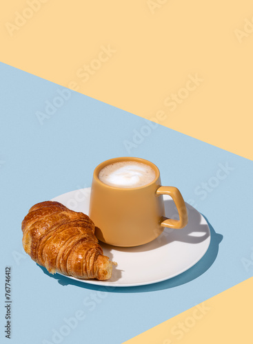 Coffee cup and fresh croissant on a white plate on a blue and yellow background.