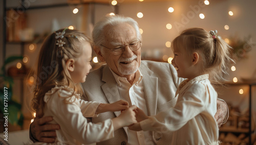 Elderly man in a light grey suit smiling, dancing with two young girls at home. The grandpa is holding the hands of his beautiful cheerful granddaughter and cute blonde doll daughter